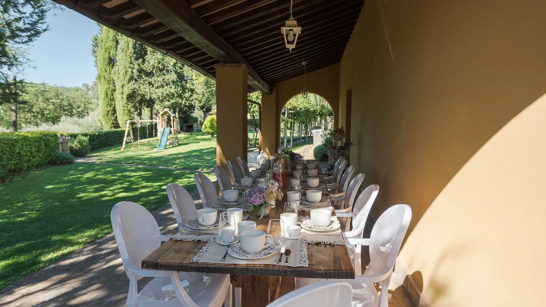 Un comedor exterior cubierto con una mesa larga preparada junto a un jardín.