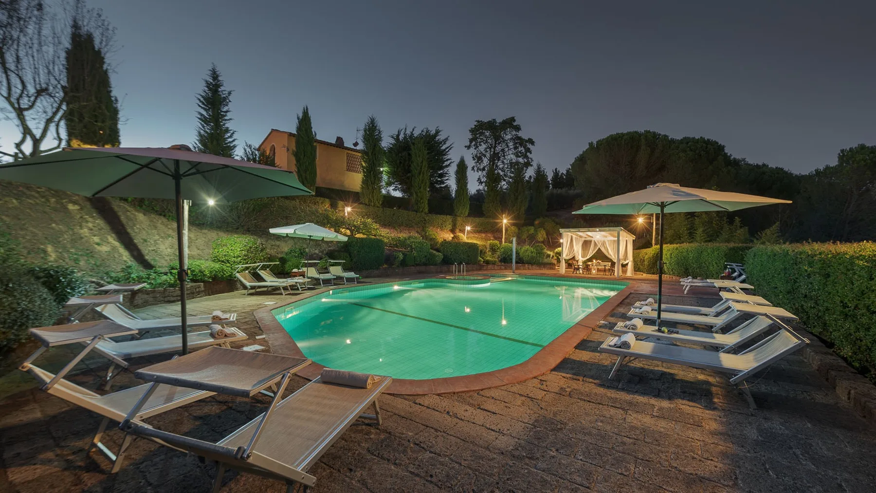 Una piscina exterior iluminada está rodeada de tumbonas y sombrillas de patio.
