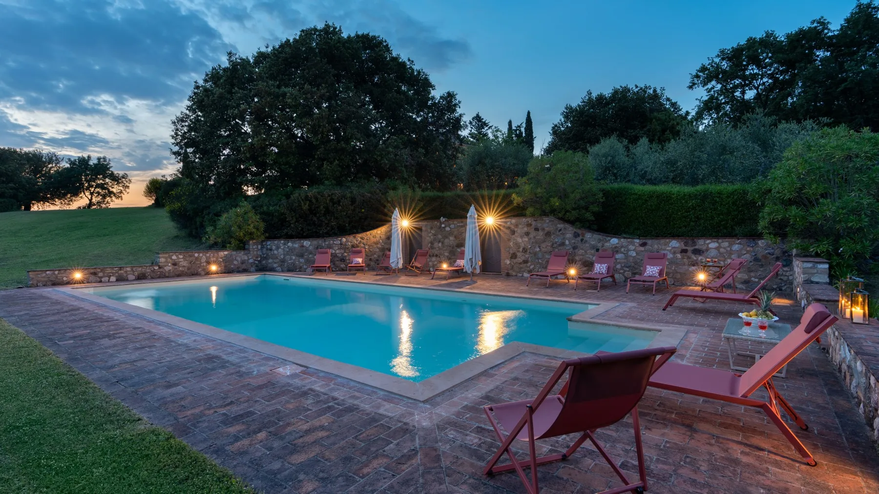 An outdoor swimming pool beside a paved patio, surrounded by trees and lounge seating.