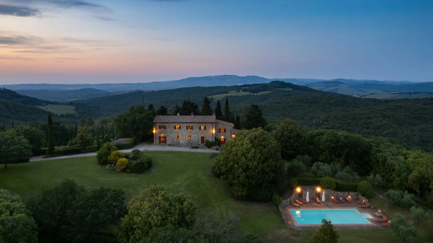 A countryside property sits among wooded hills, with a lit swimming pool below.