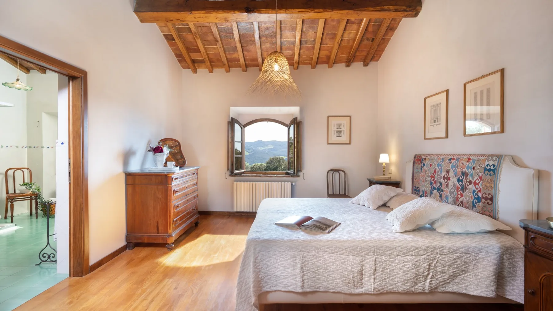 A bedroom with a large bed, wooden ceiling beams, and a window overlooking hills.