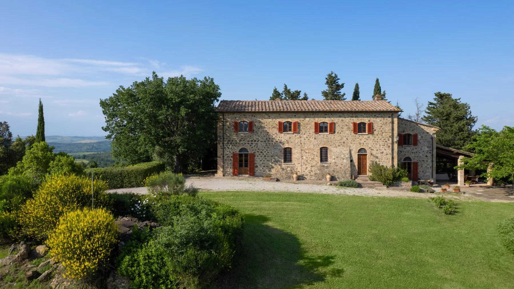 A stone house stands beyond a green lawn, with trees and hills in the distance.