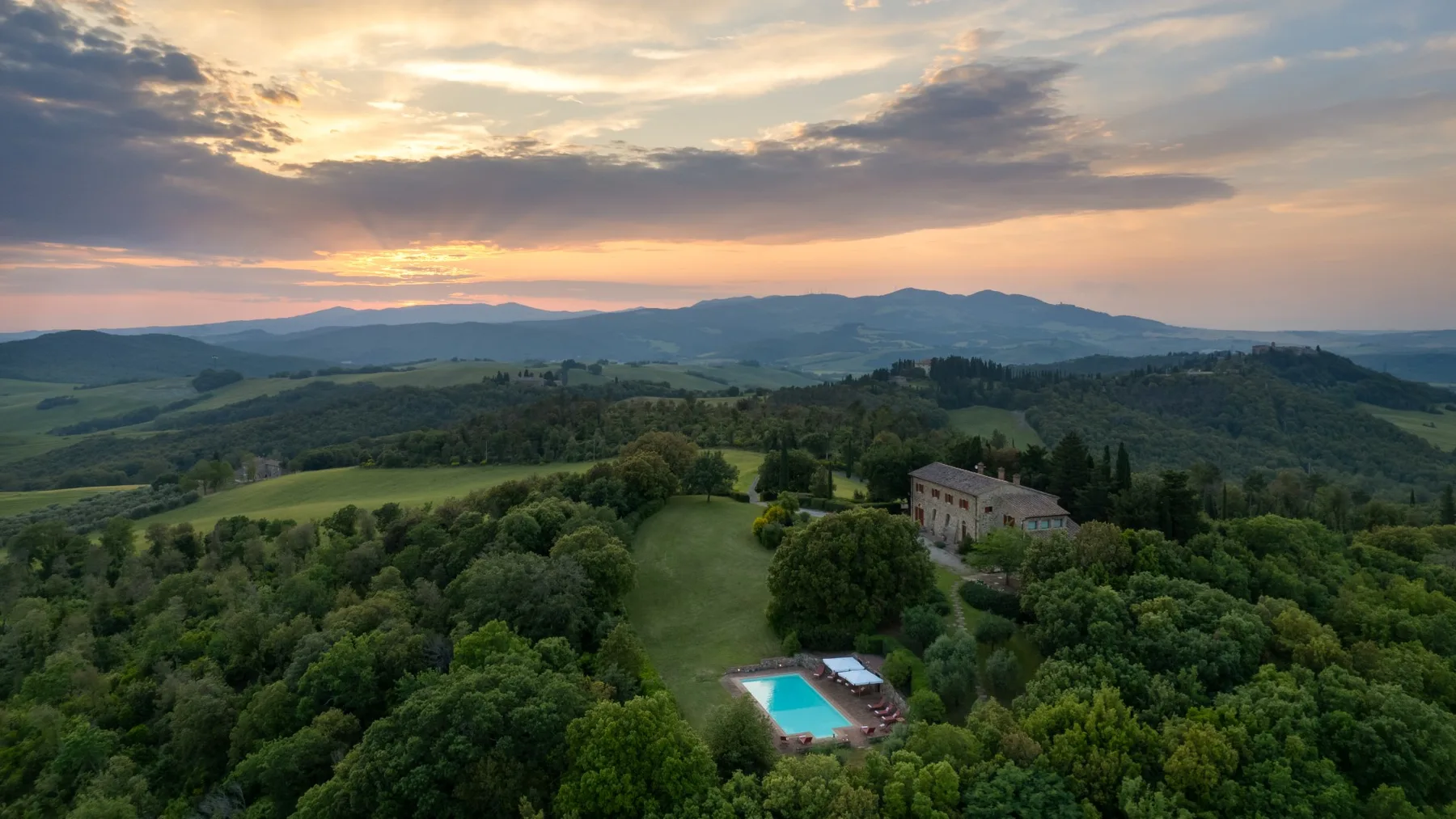 Aerial view of wooded hills and a swimming pool beneath a colorful sunset sky.