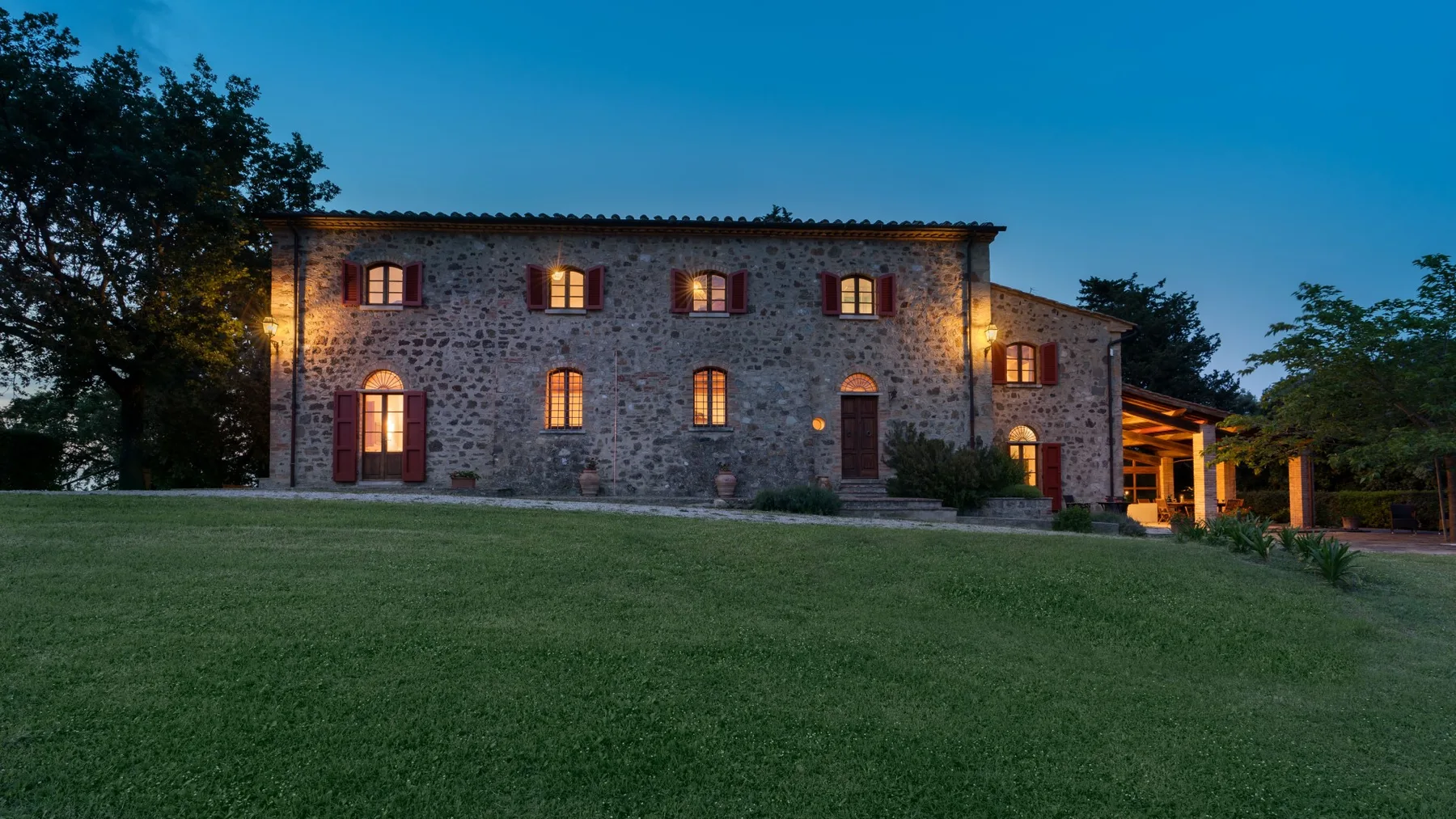 A stone house stands beyond a broad lawn at dusk, with warm light glowing from its windows.