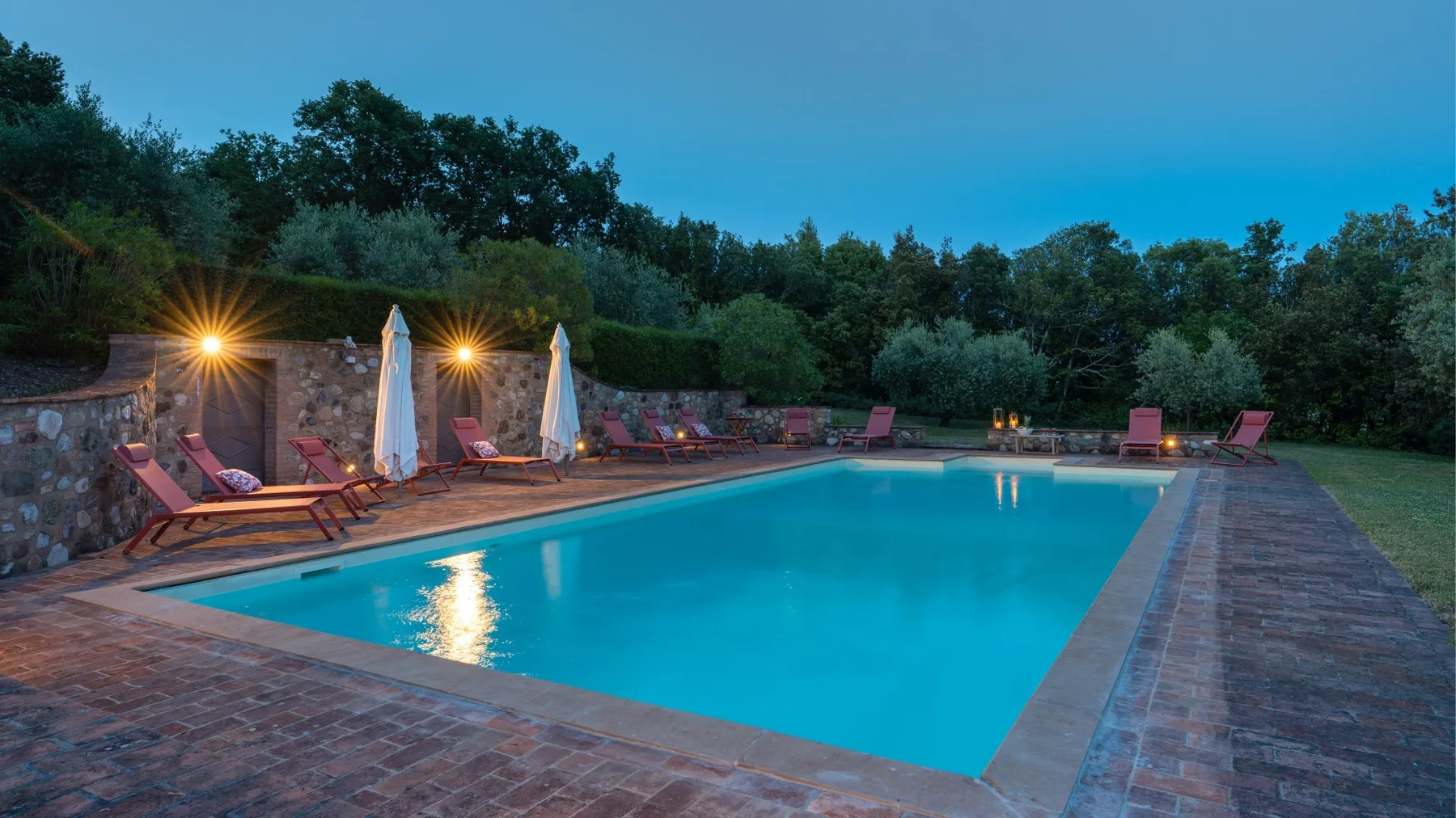 An outdoor swimming pool is surrounded by loungers and trees at dusk.