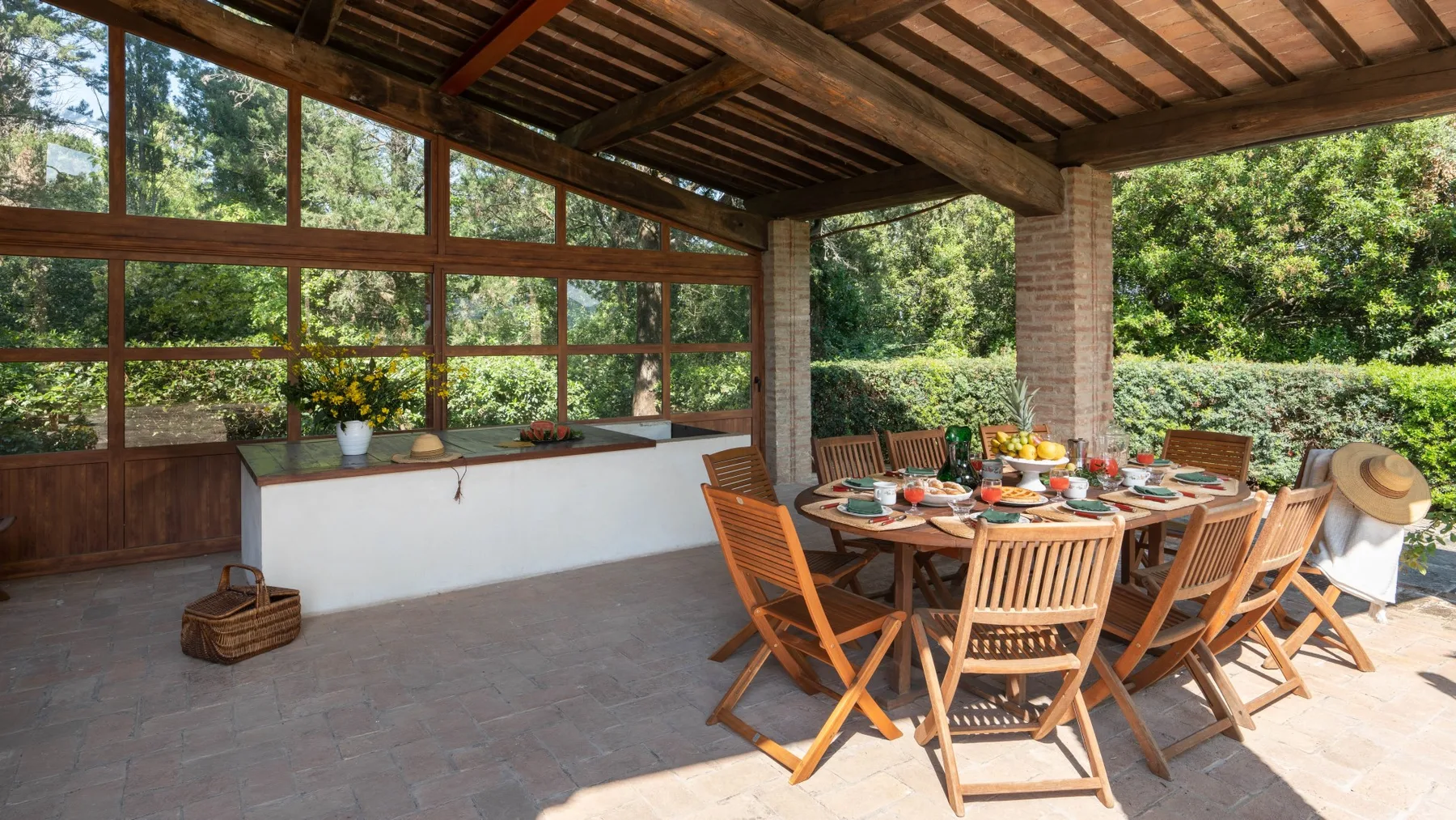 A covered outdoor dining area with a wooden table set beside large windows.