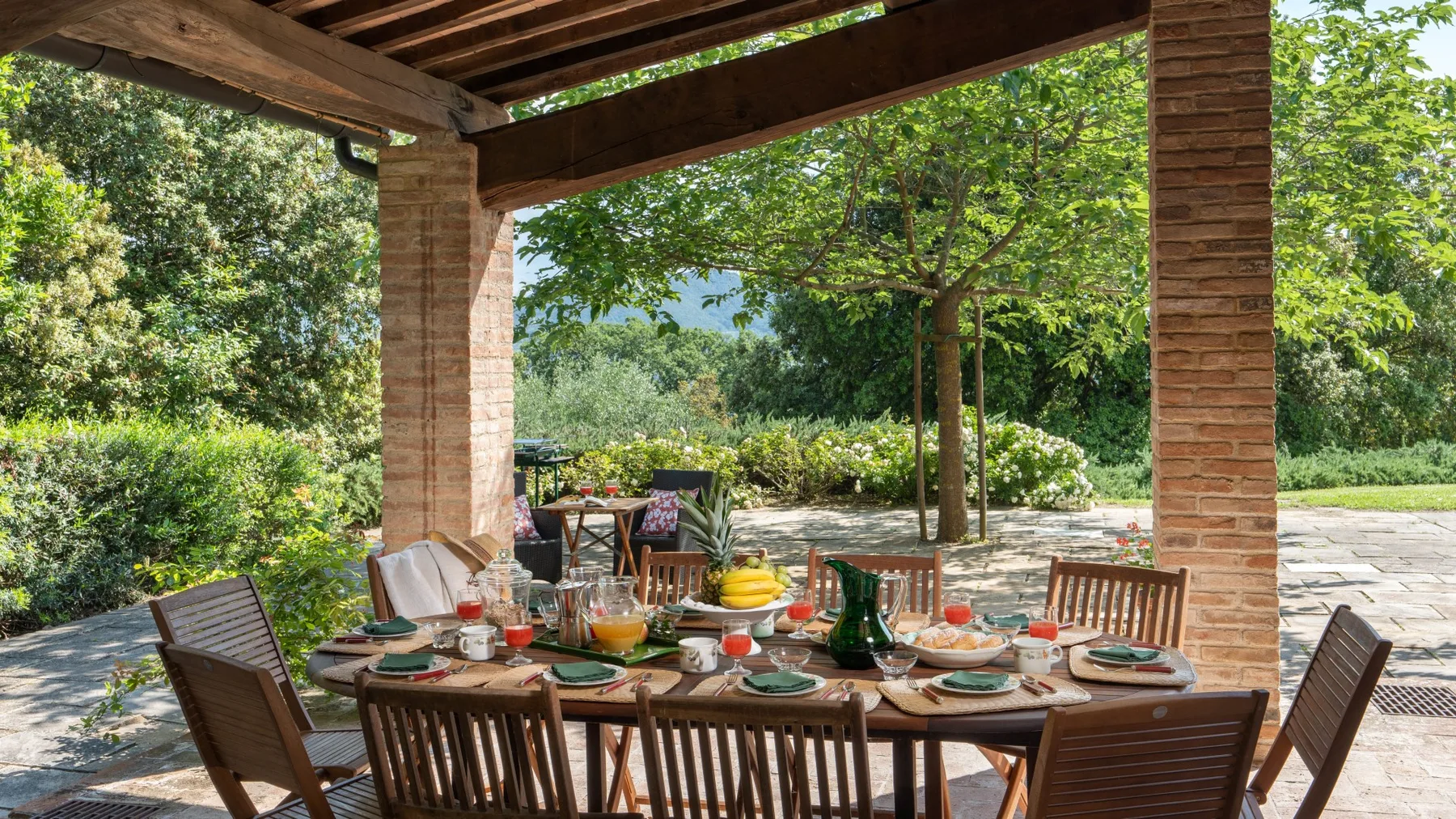 A covered outdoor dining area with a long table set among trees and greenery.