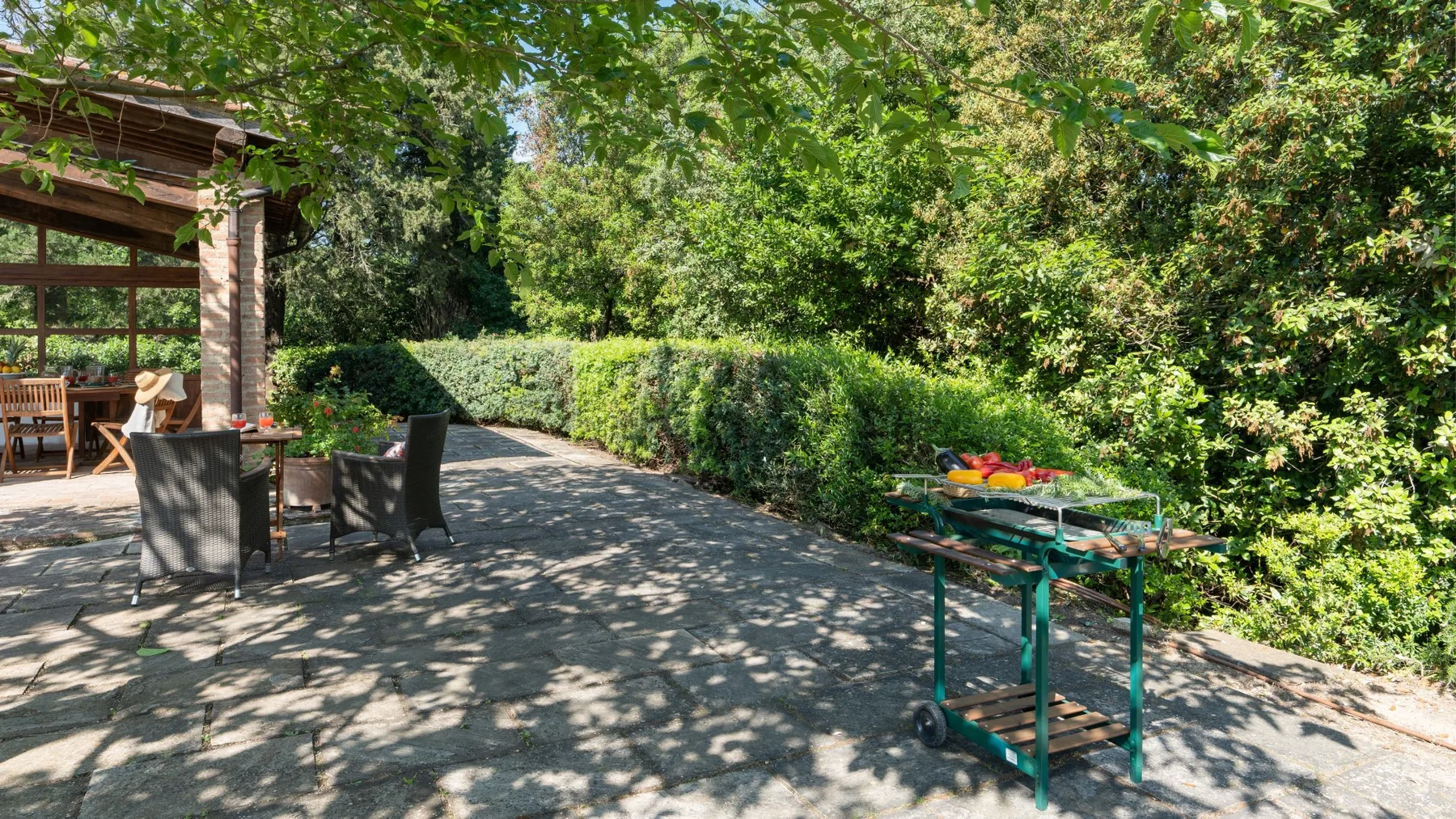 A shaded outdoor patio with seating beside dense greenery and a garden table.
