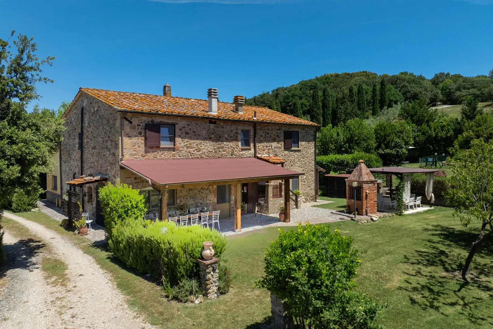 A stone country house with a covered patio overlooks a garden and wooded hills.
