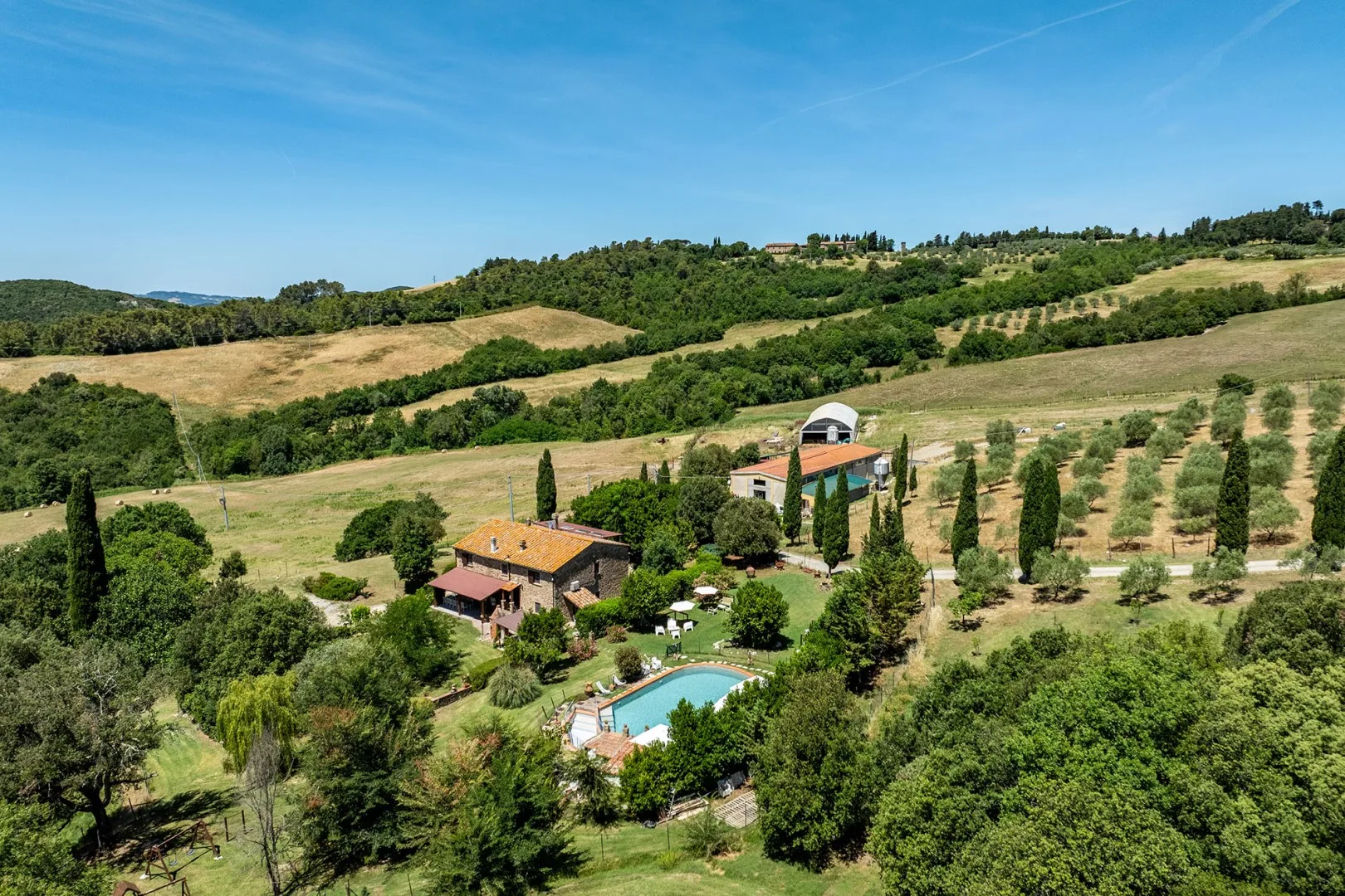 A countryside property sits among rolling hills, with a pool and rows of trees.