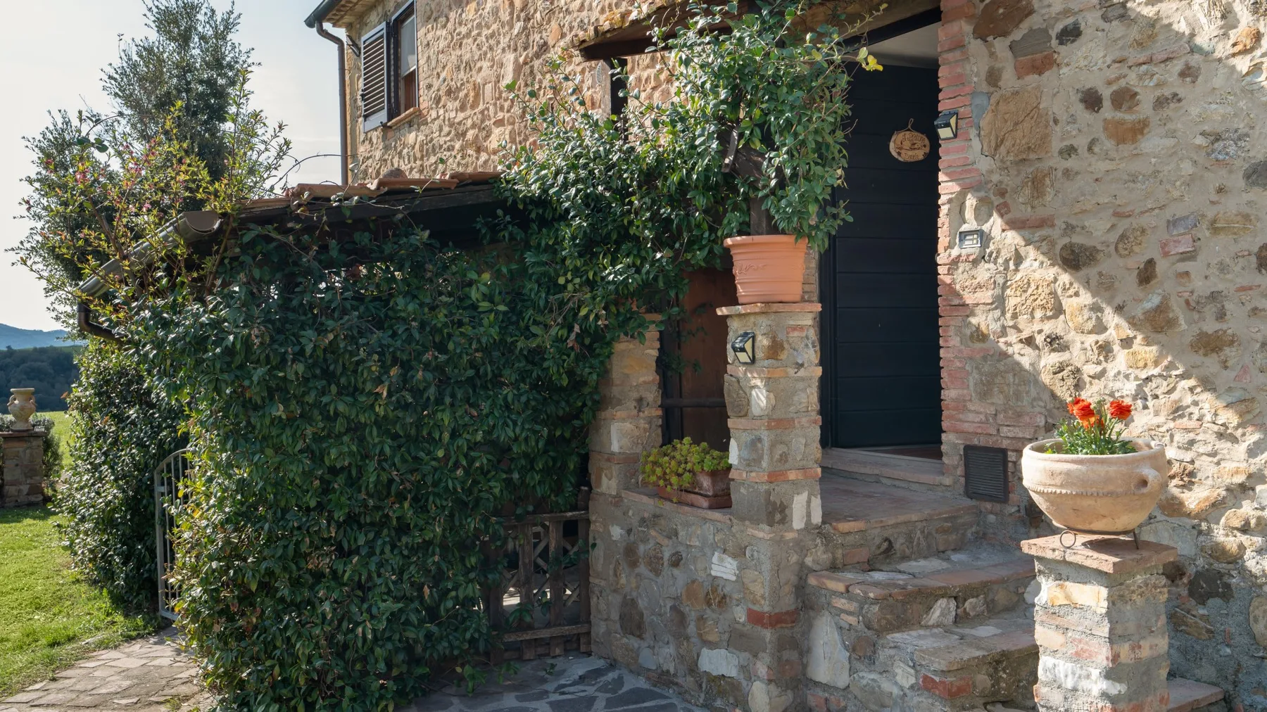 A stone house entrance with steps, potted flowers, and climbing greenery.