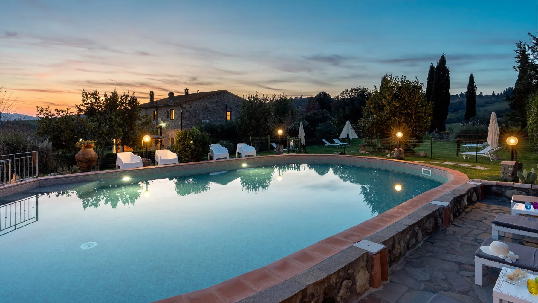 An outdoor swimming pool at dusk, with a stone building and trees beyond.