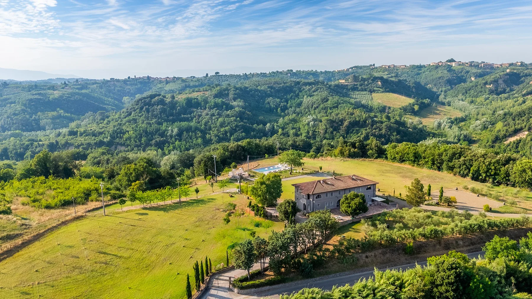 Una casa rural se encuentra entre campos verdes y colinas bajo un cielo parcialmente nublado.