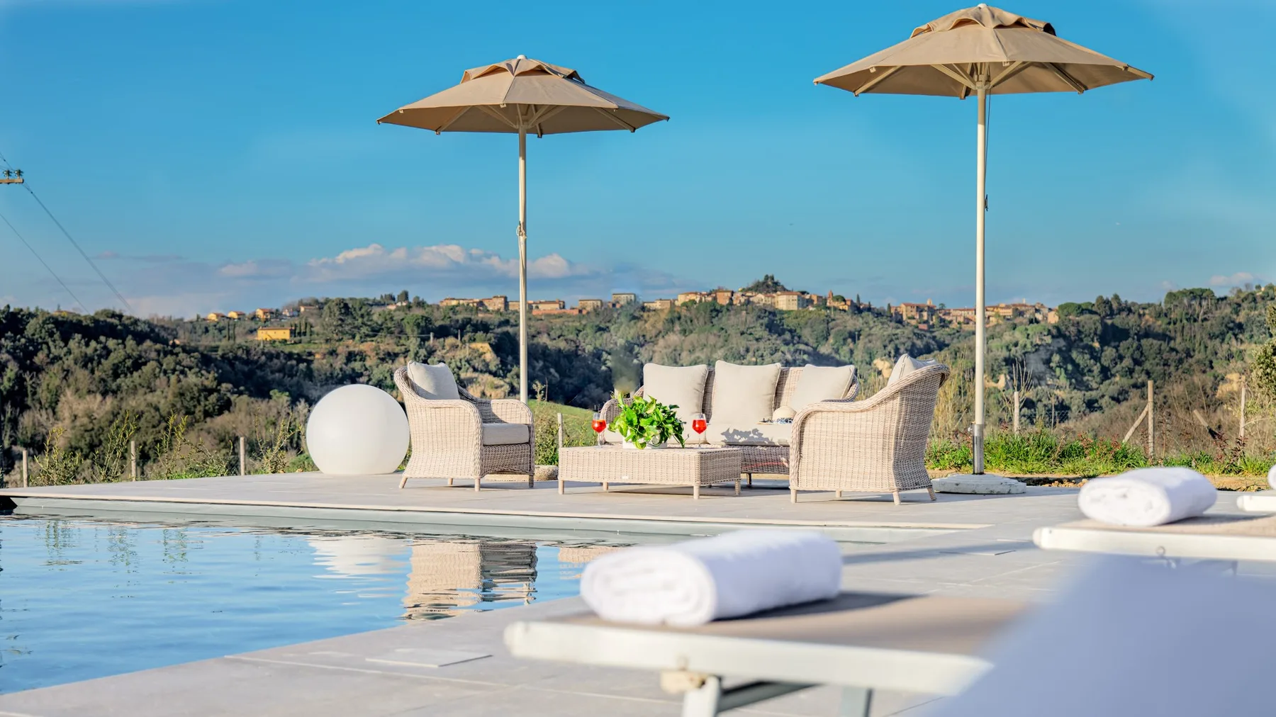 Un patio junto a la piscina con asientos acolchados y sombrillas da a un paisaje arbolado.