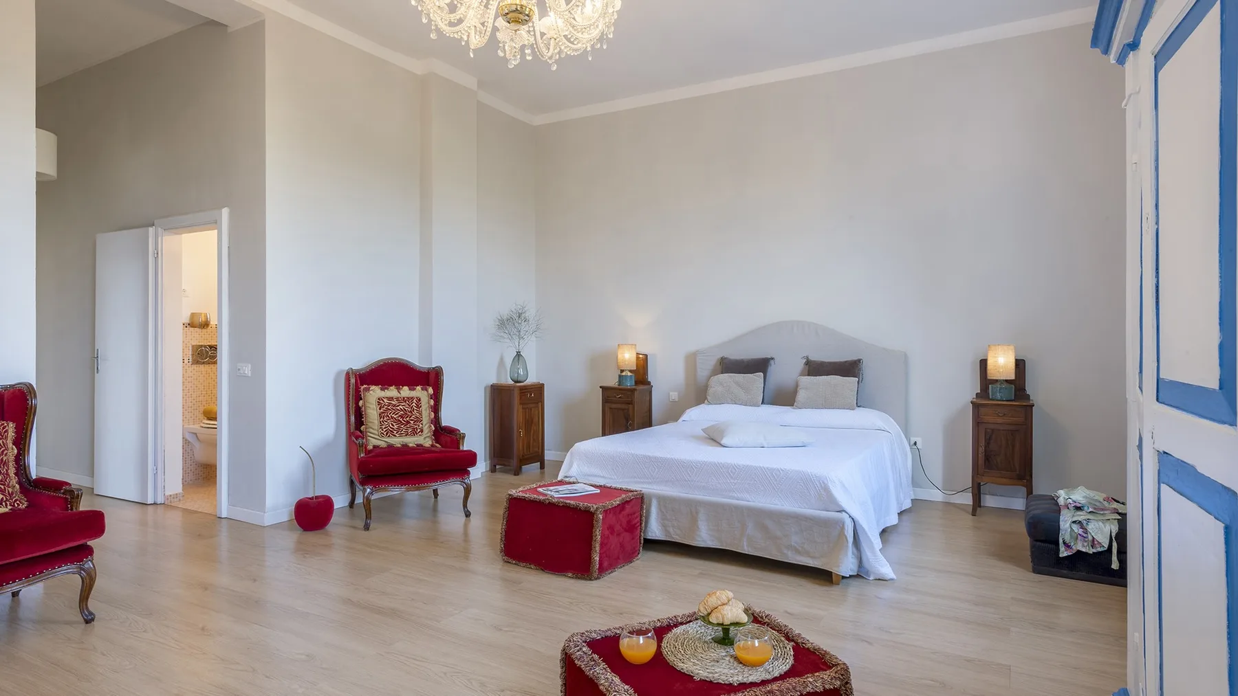 A bright bedroom with a large bed, red seating, and a chandelier.