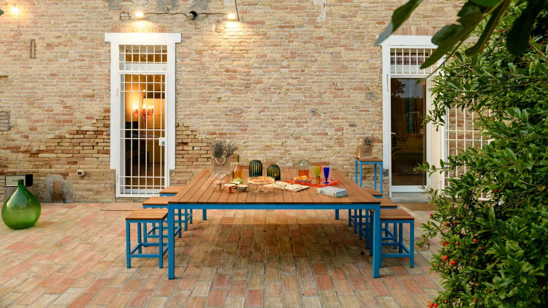 An outdoor dining area with a long table and blue seating beside a brick wall.