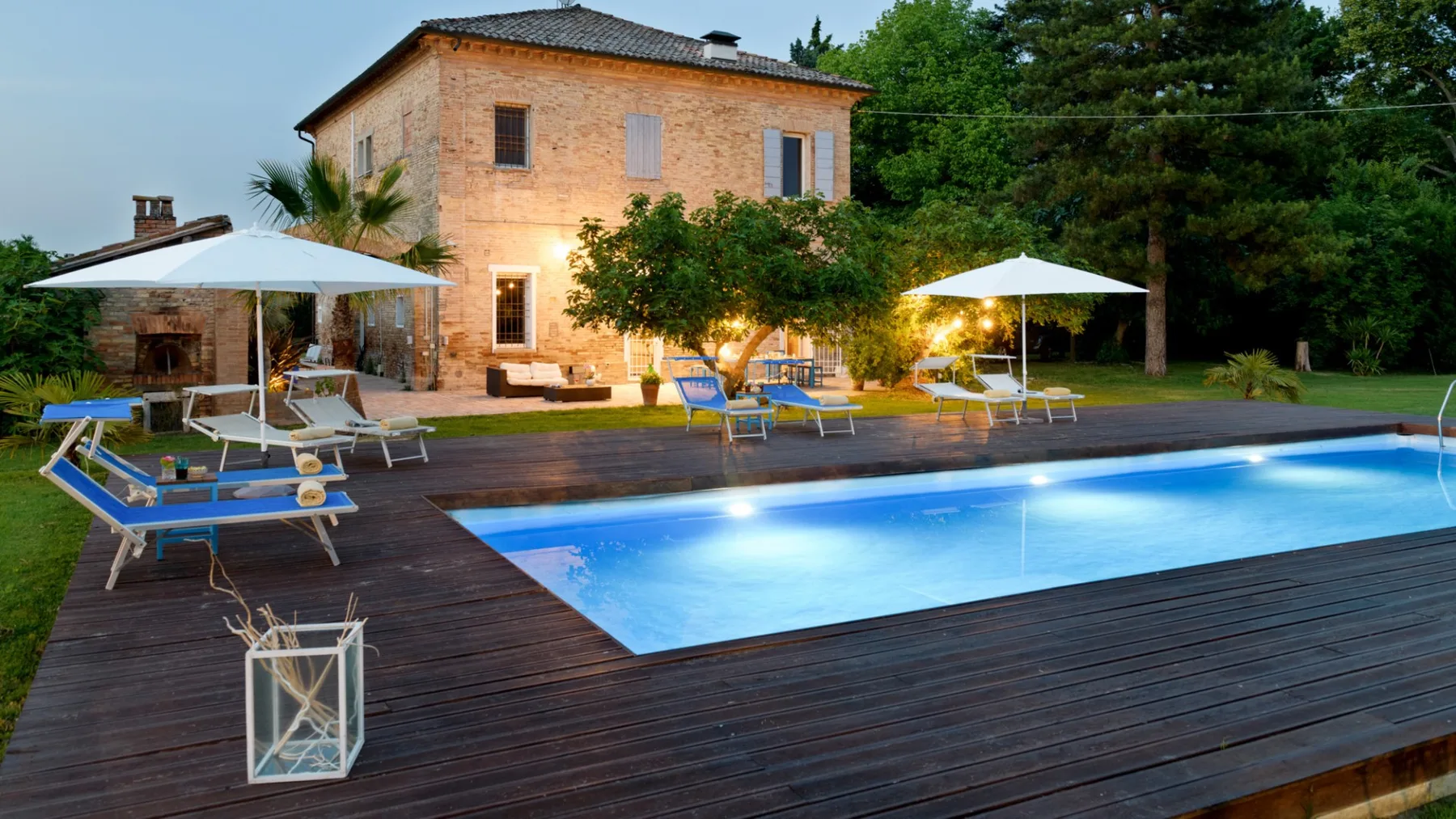 A swimming pool and wooden deck sit beside a two-story stone house.