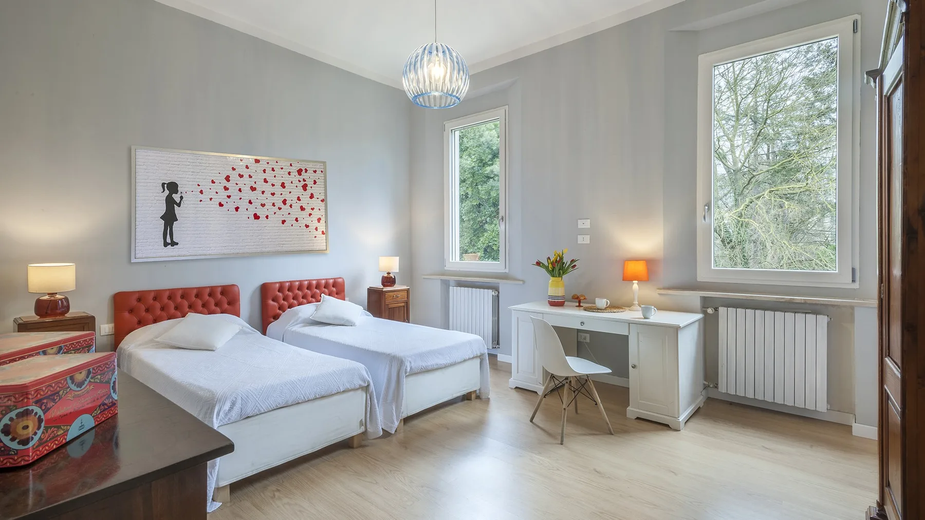 A bright bedroom with two beds and a desk beside the windows.
