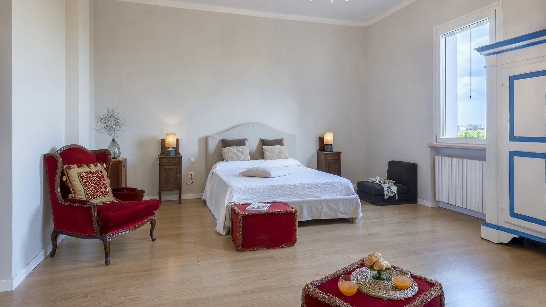 A bedroom with a bed, red accent chair, and a large window.