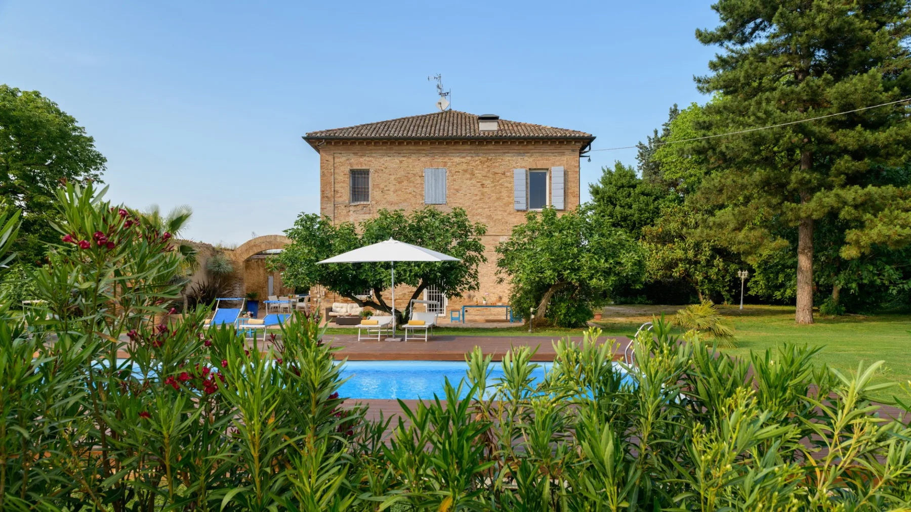 An outdoor swimming pool sits among greenery in front of a two-story house.