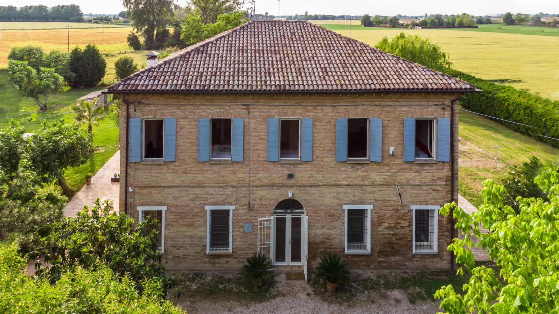 A two-story brick house stands amid green trees and open fields.
