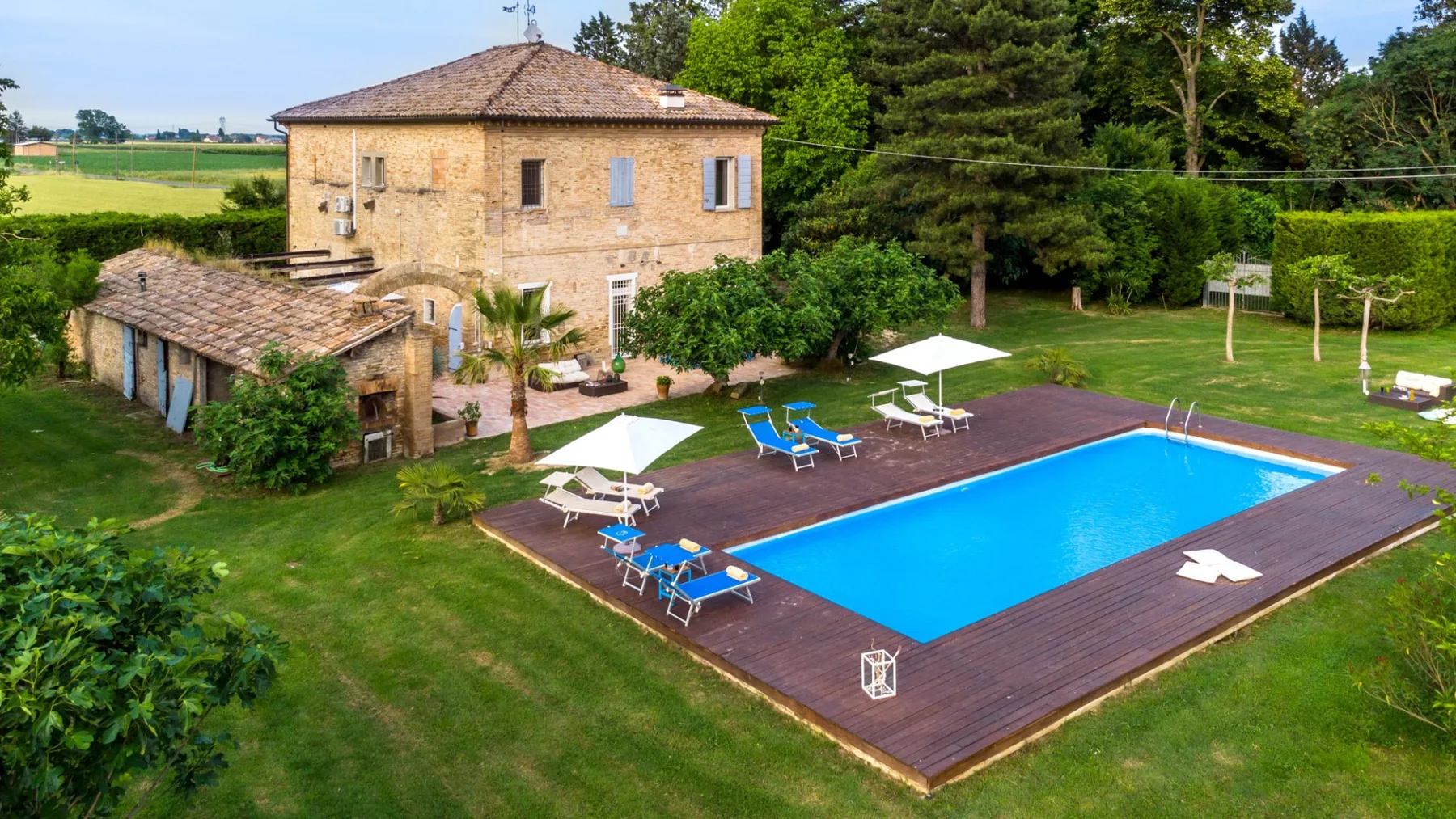 A countryside house overlooks a swimming pool and a lawn with outdoor seating.