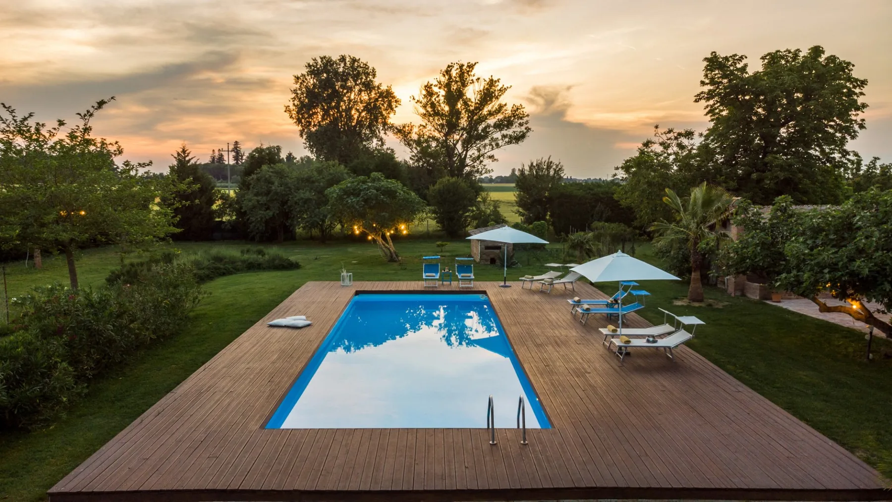 An outdoor swimming pool sits on a wooden deck amid trees at sunset.