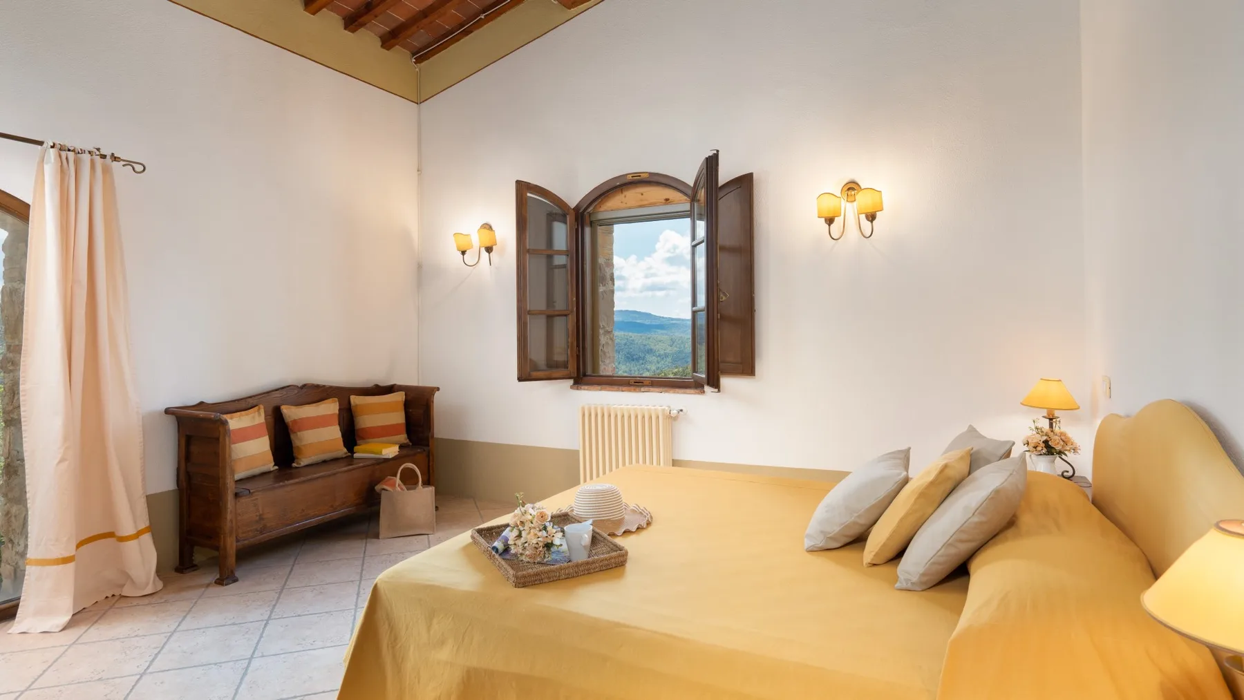 Un dormitorio con una cama amarilla, un banco de madera y una ventana abierta con vistas a colinas.