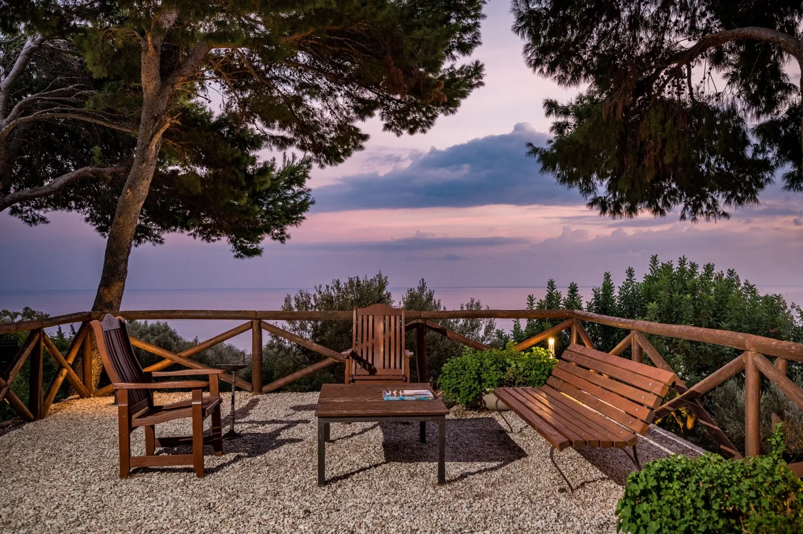 Una terraza de grava con asientos de madera da al mar al atardecer.