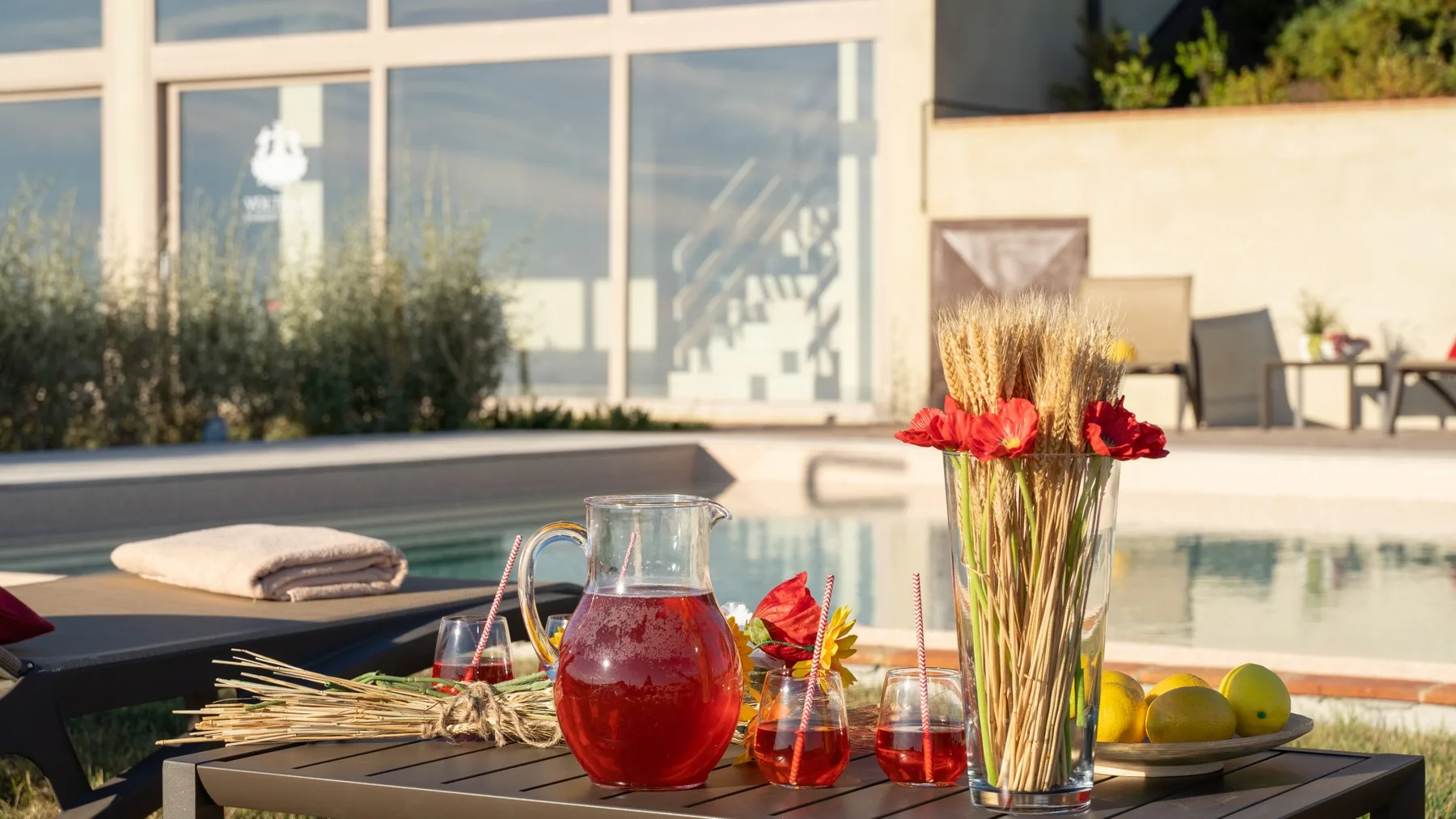 A poolside table displays a pitcher of red drink, glasses, fruit, and flowers.