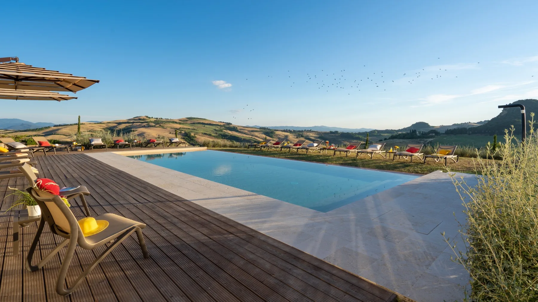 An outdoor swimming pool and wooden deck overlook rolling hills under a clear blue sky.