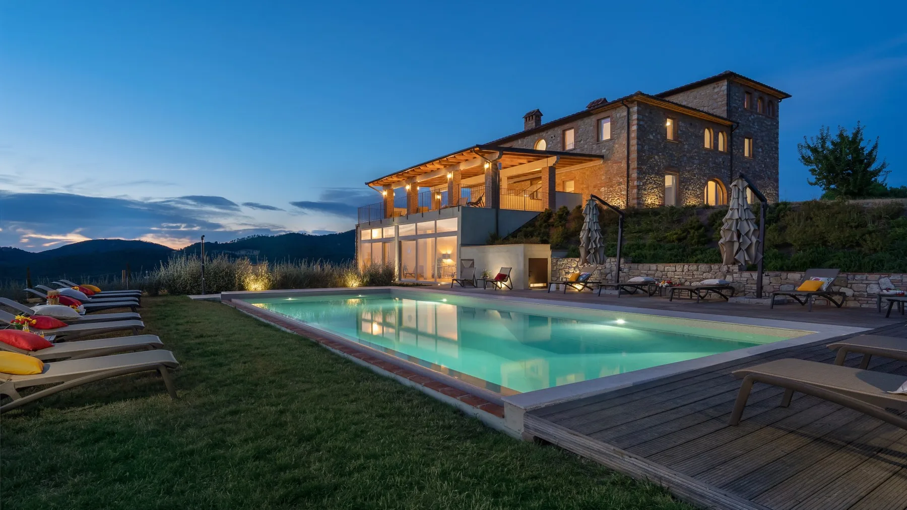 A lit stone house overlooks a swimming pool and outdoor seating at dusk.