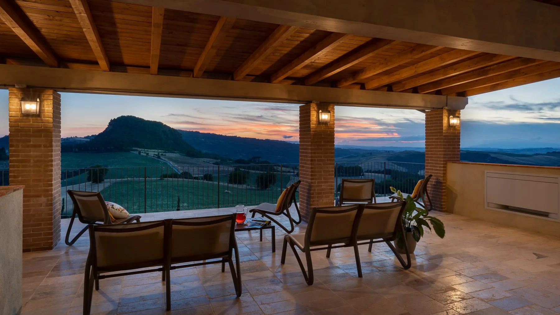 A covered outdoor seating area overlooks a hilly landscape at sunset.
