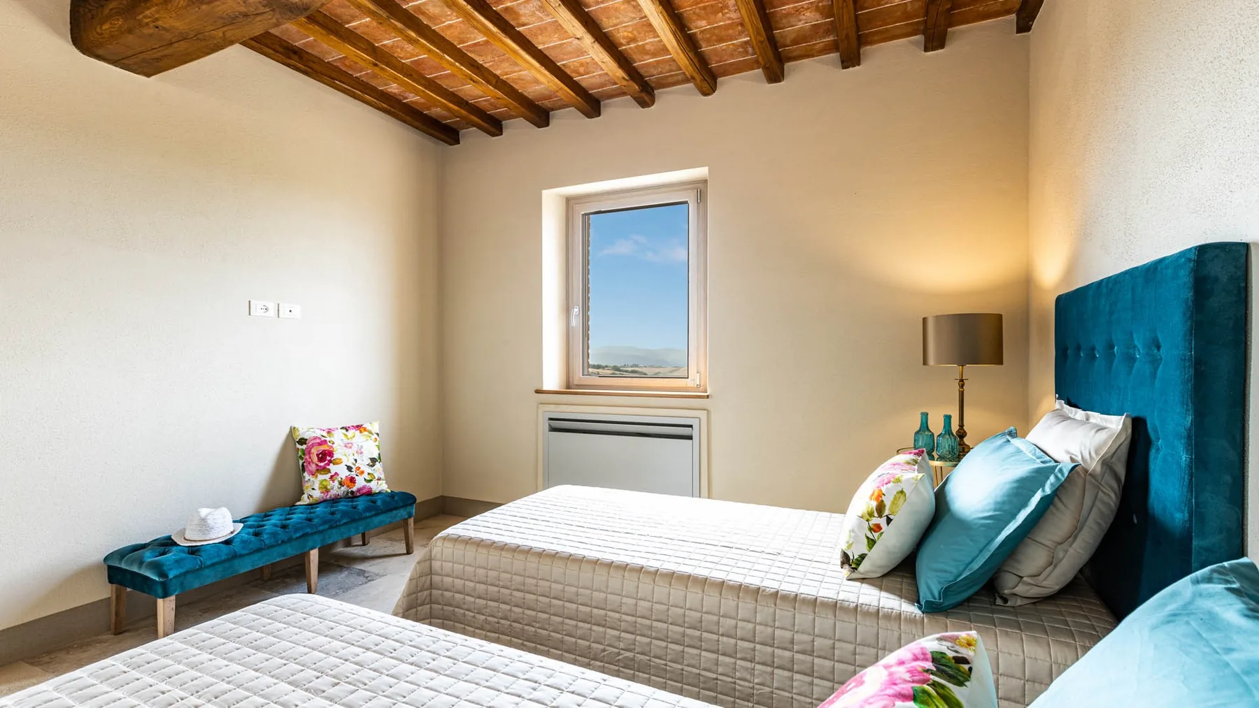 A bedroom with a bed, teal headboard, and a window beneath exposed wooden beams.