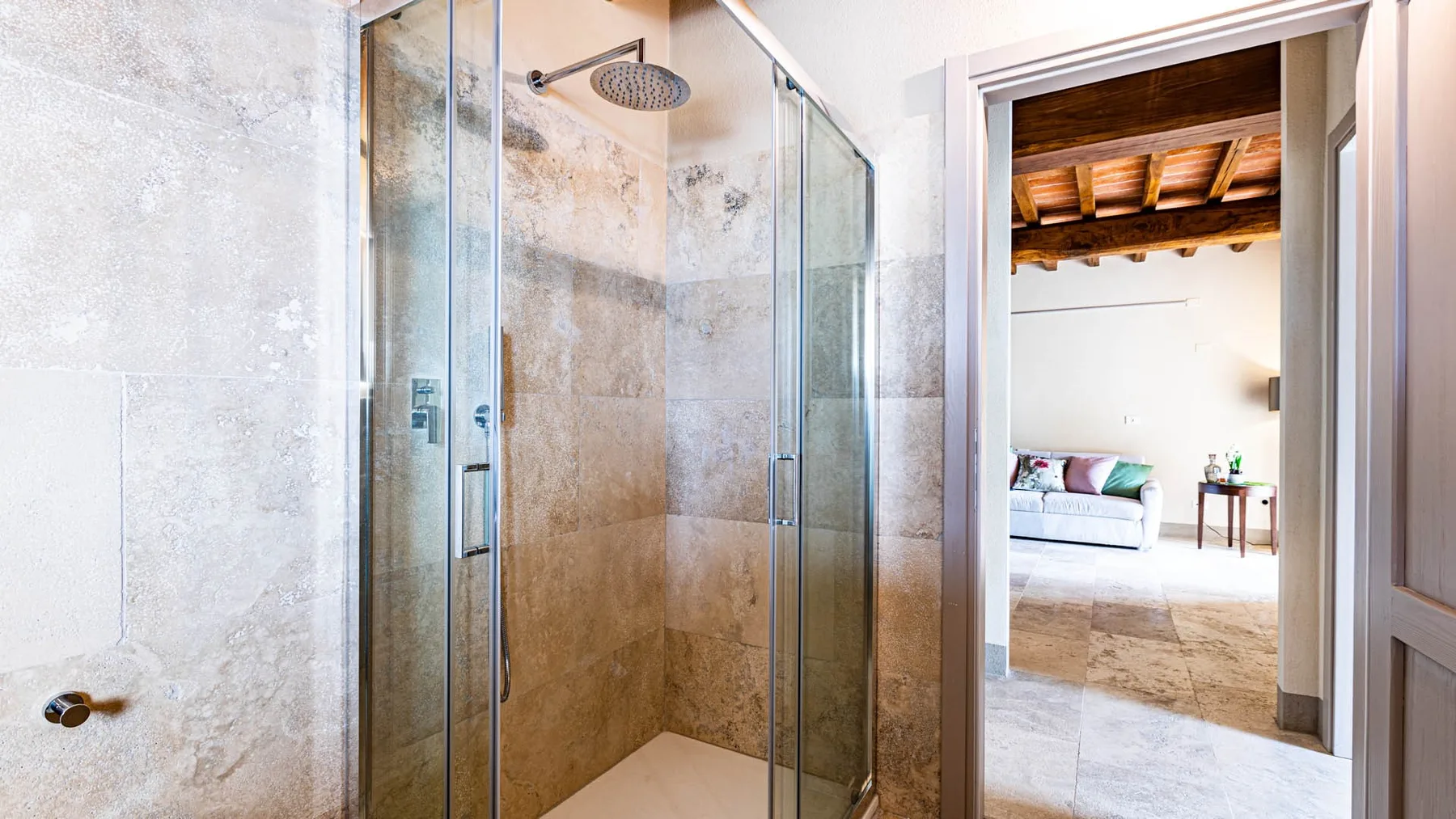 A tiled bathroom with a glass shower and an open doorway to a bright room.