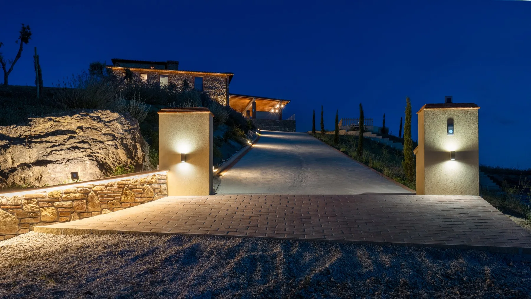 A lit driveway leads uphill toward a modern house at dusk.