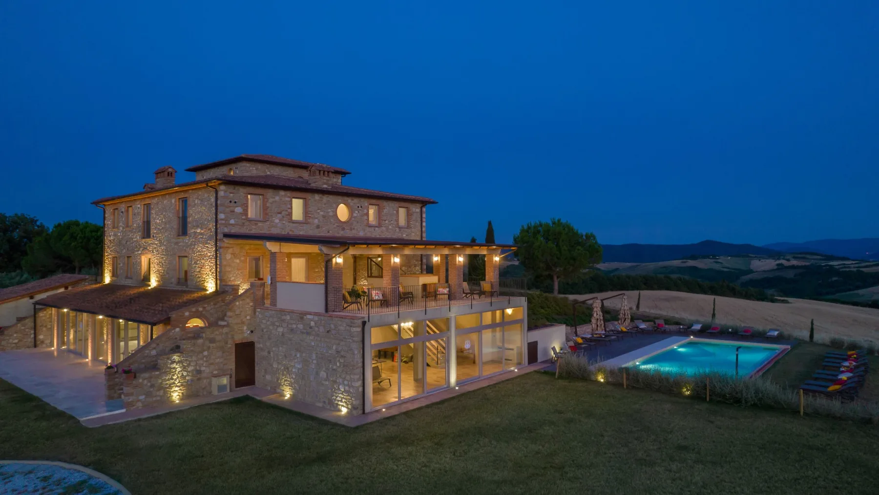 A stone building with illuminated windows stands beside an outdoor swimming pool at dusk.