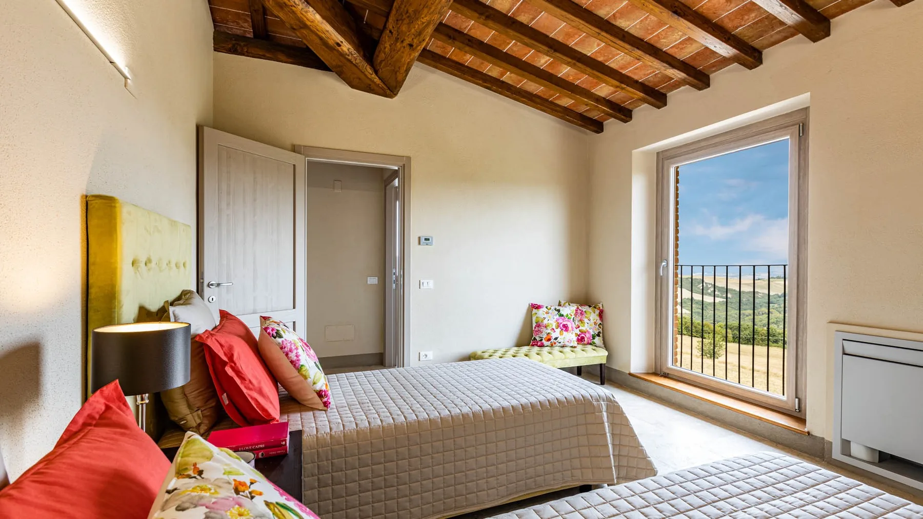 A bedroom with beds, colorful pillows, and a large window opening onto a balcony.