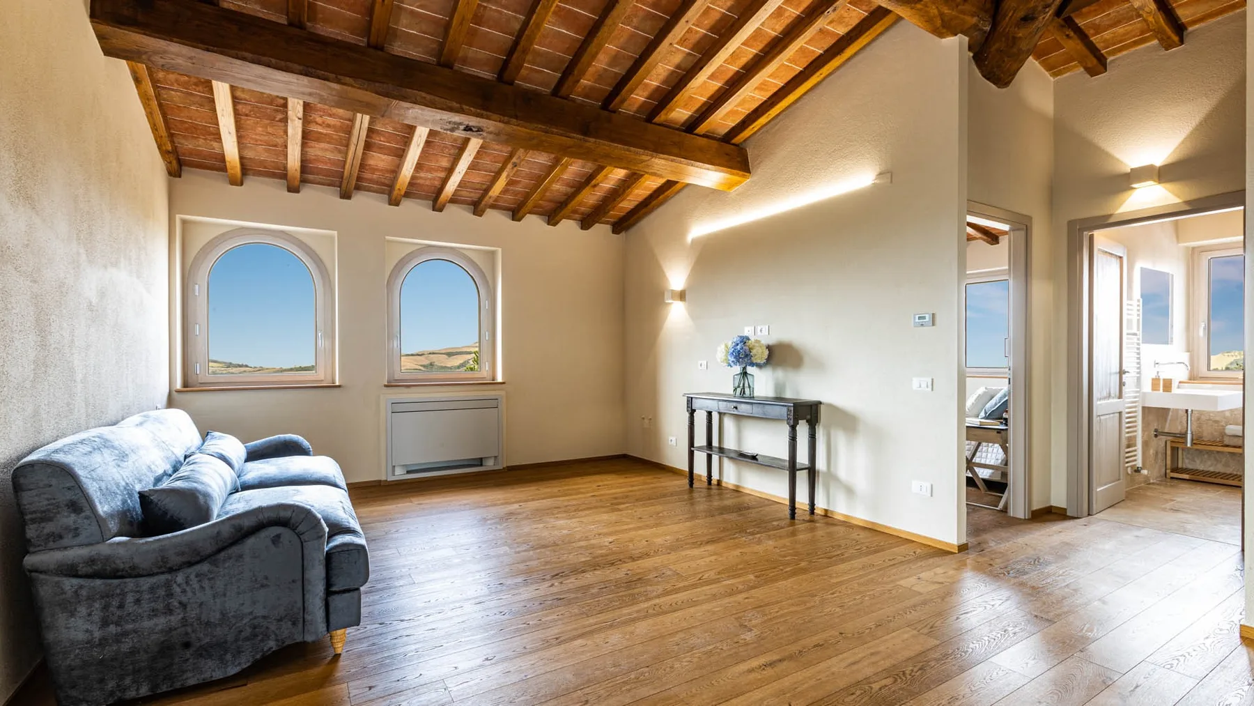 A bright living area with a gray sofa, wooden floor, and exposed beams.