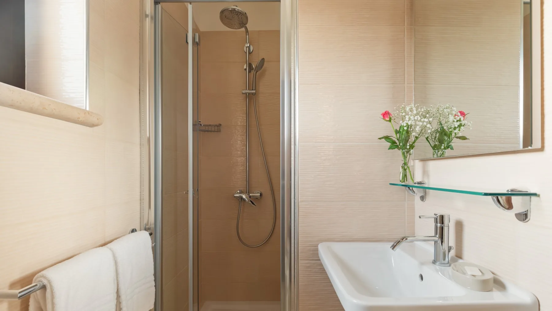 A bathroom with a glass shower, sink, and flowers on a shelf.