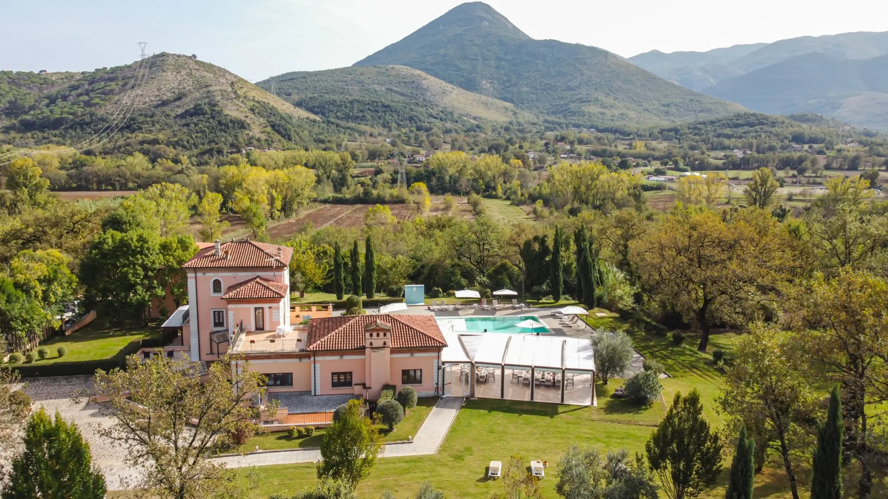 A large house and covered pool sit in a green landscape with hills behind.