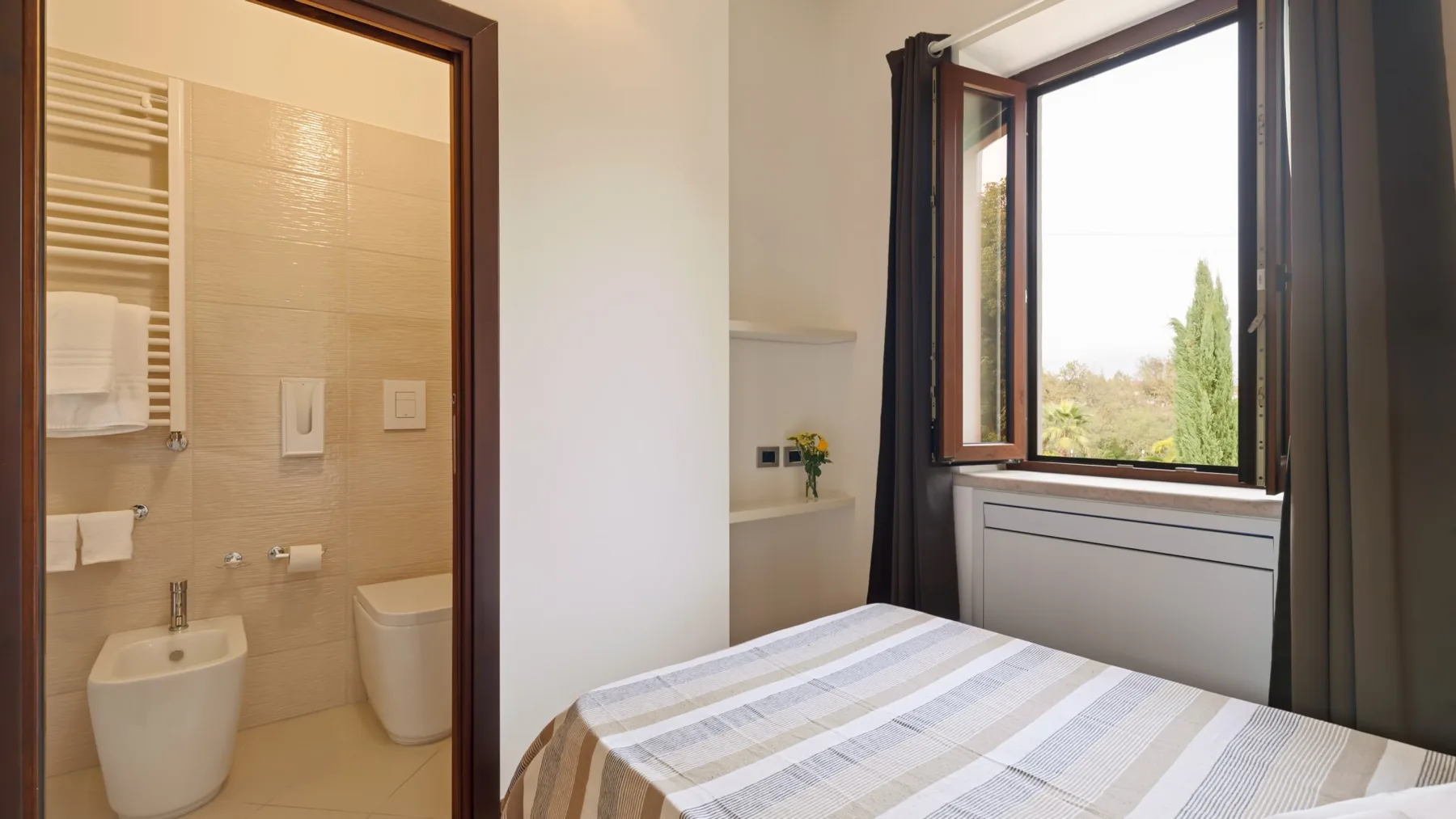 A bedroom with a neatly made bed beside an open window and an adjoining bathroom.