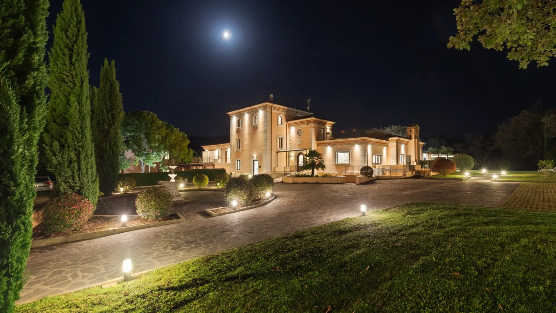 A warmly lit villa stands beside a paved drive and landscaped grounds at night.