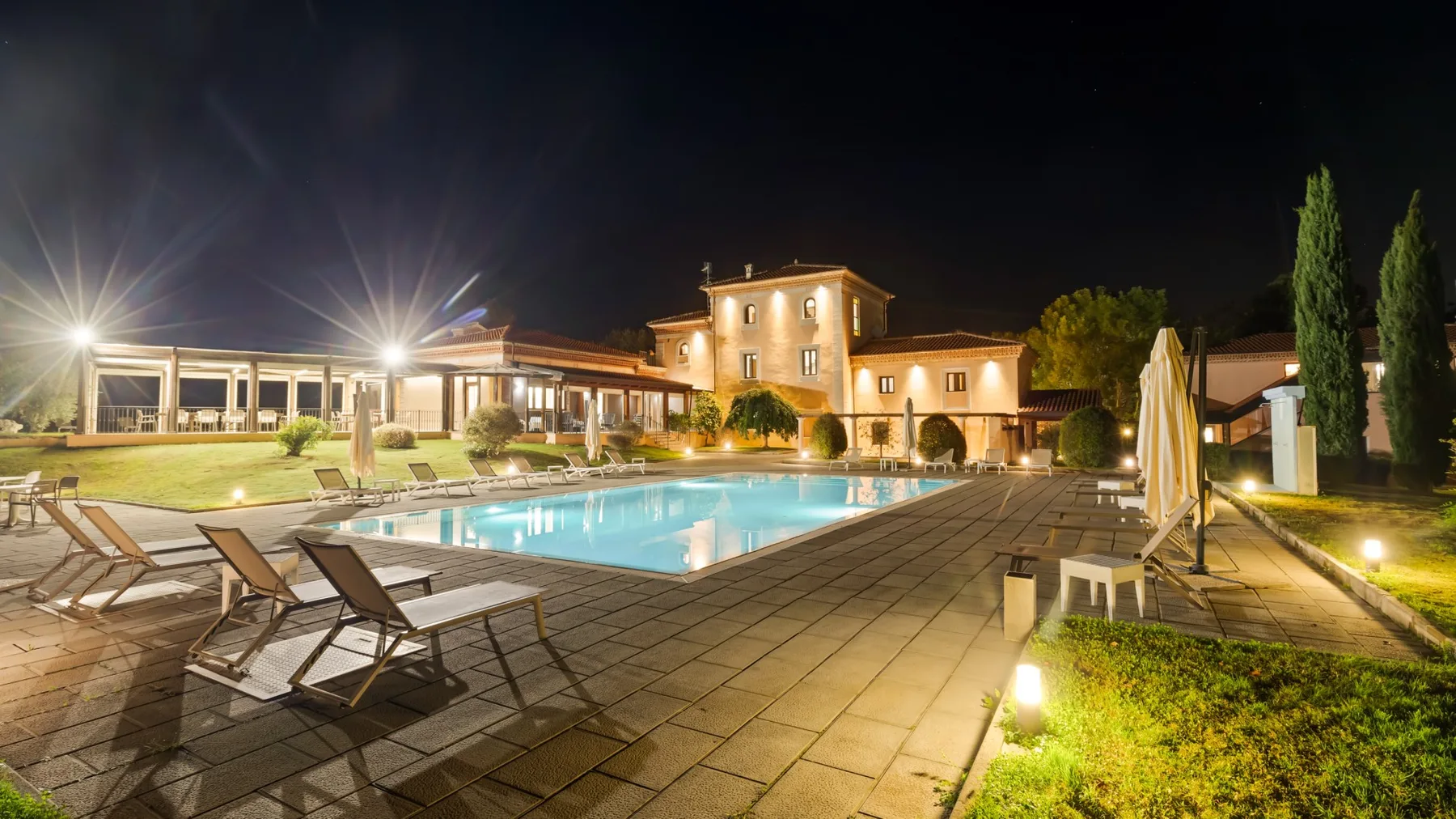 A lit outdoor swimming pool and paved patio sit beside a large building at night.