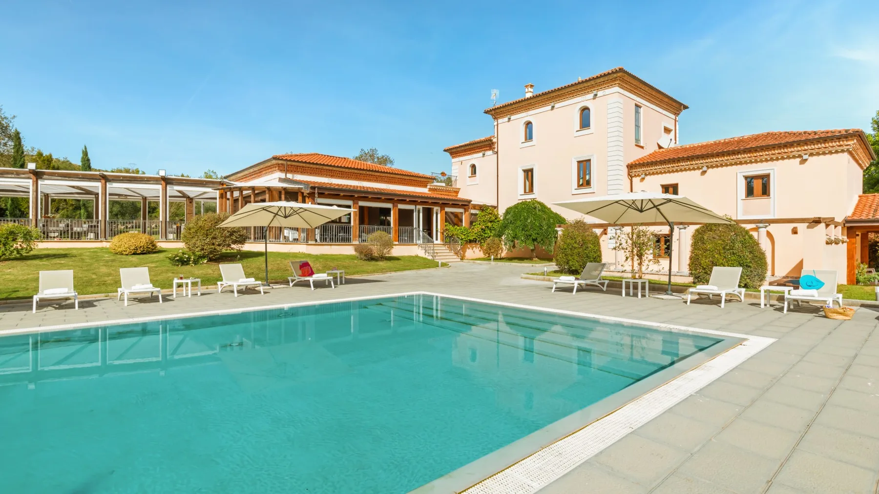 An outdoor swimming pool beside a pale building and landscaped grounds.