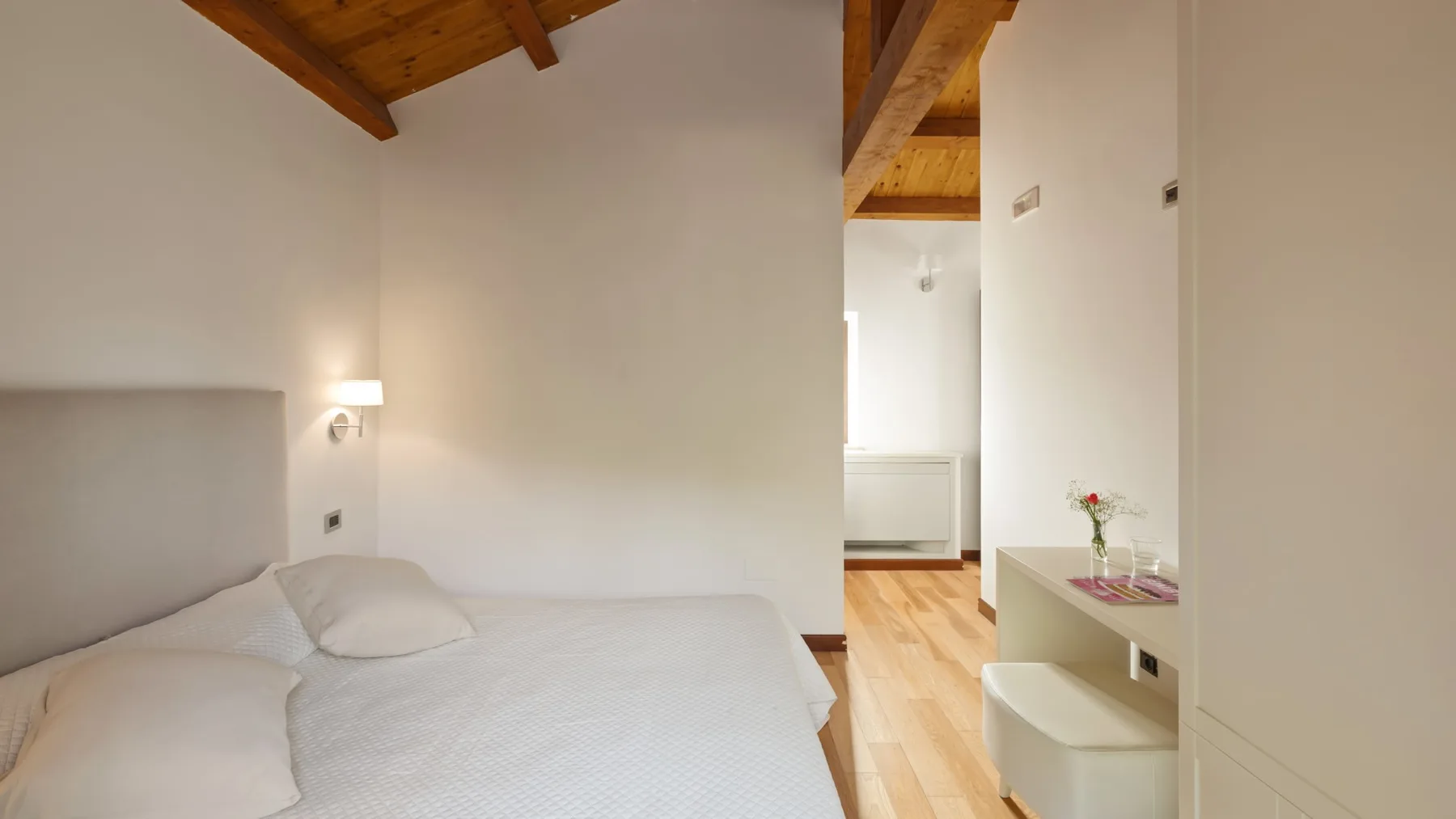 A bright bedroom with a bed, wooden ceiling beams, and a small display shelf.
