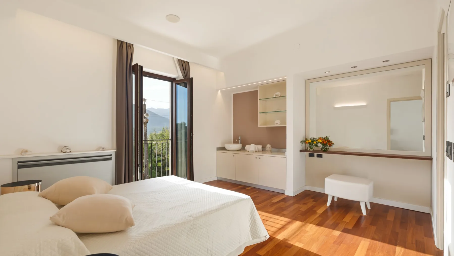 A bright bedroom with a bed, an open balcony door, and a small vanity area.