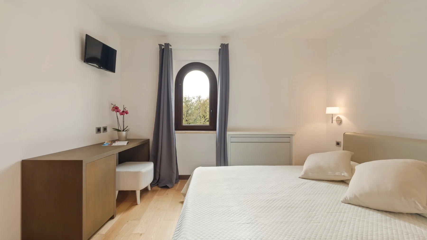 A bright bedroom with a bed, a wooden dresser, and an arched window.