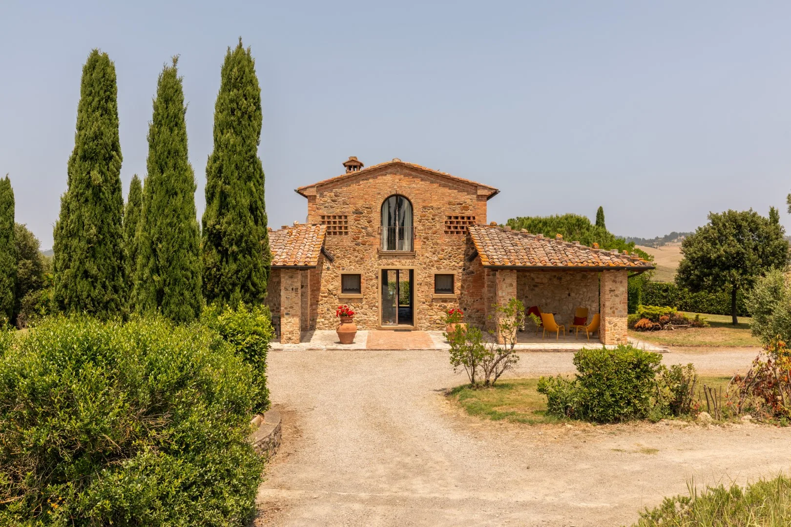 A brick country house stands at the end of a gravel drive, surrounded by trees and shrubs.