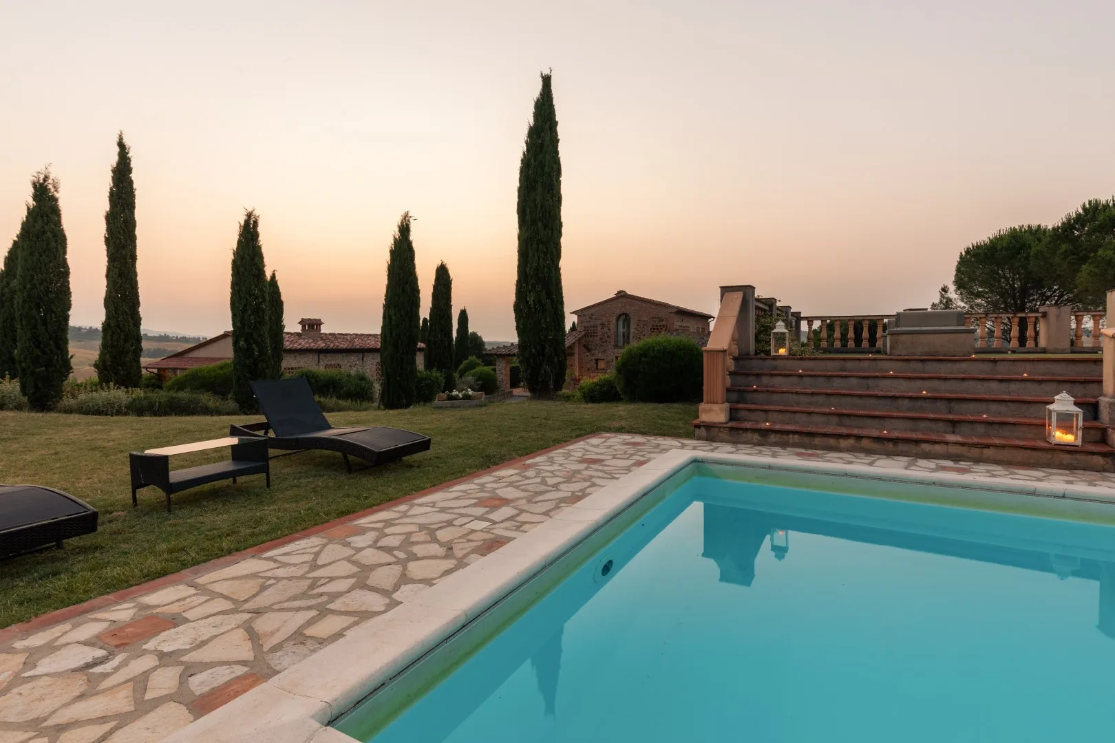 An outdoor swimming pool beside a stone patio and landscaped garden at sunset.